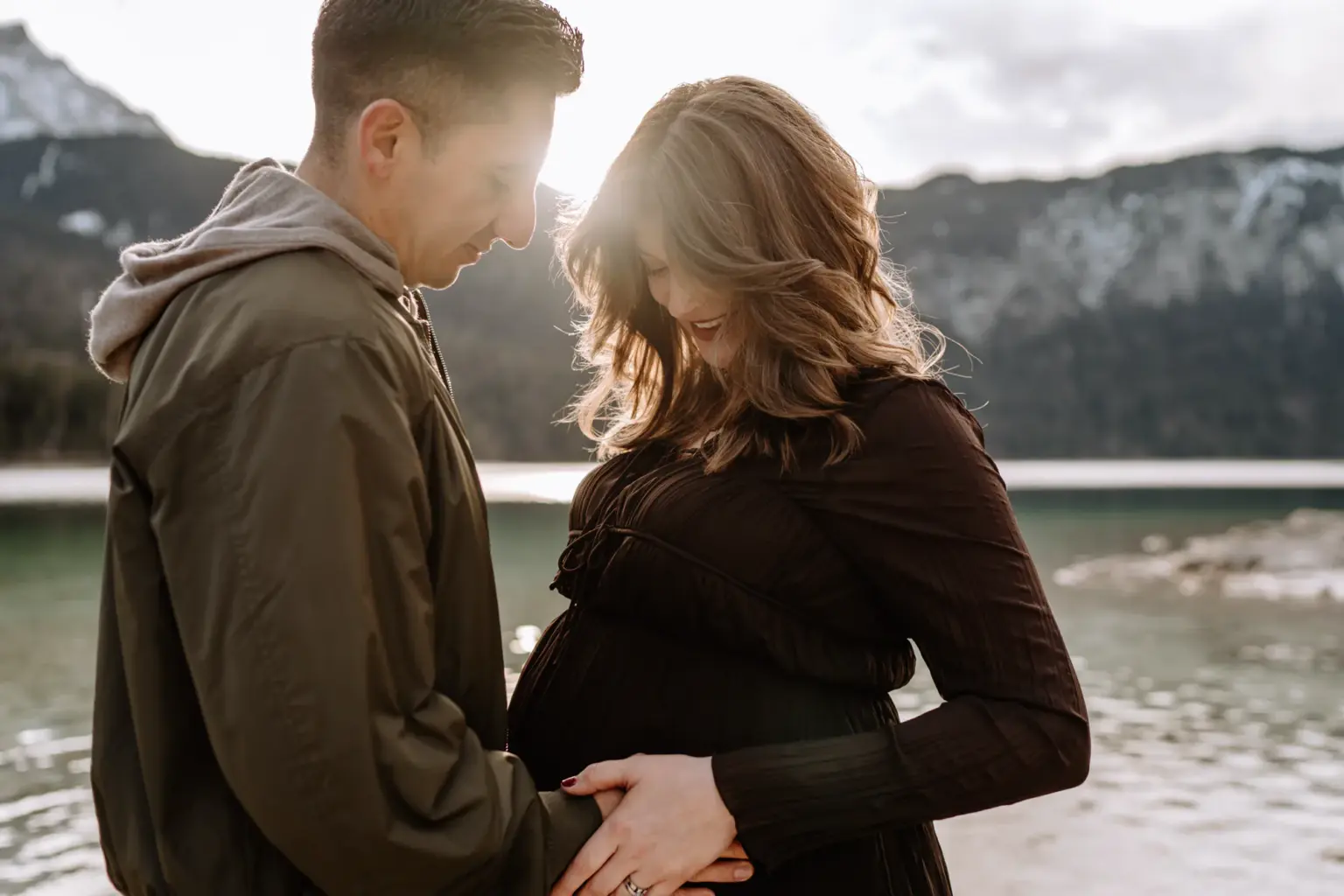 Paar hält den Babybauch gemeinsam, Eibsee Hintergrund, natürliche Nähe