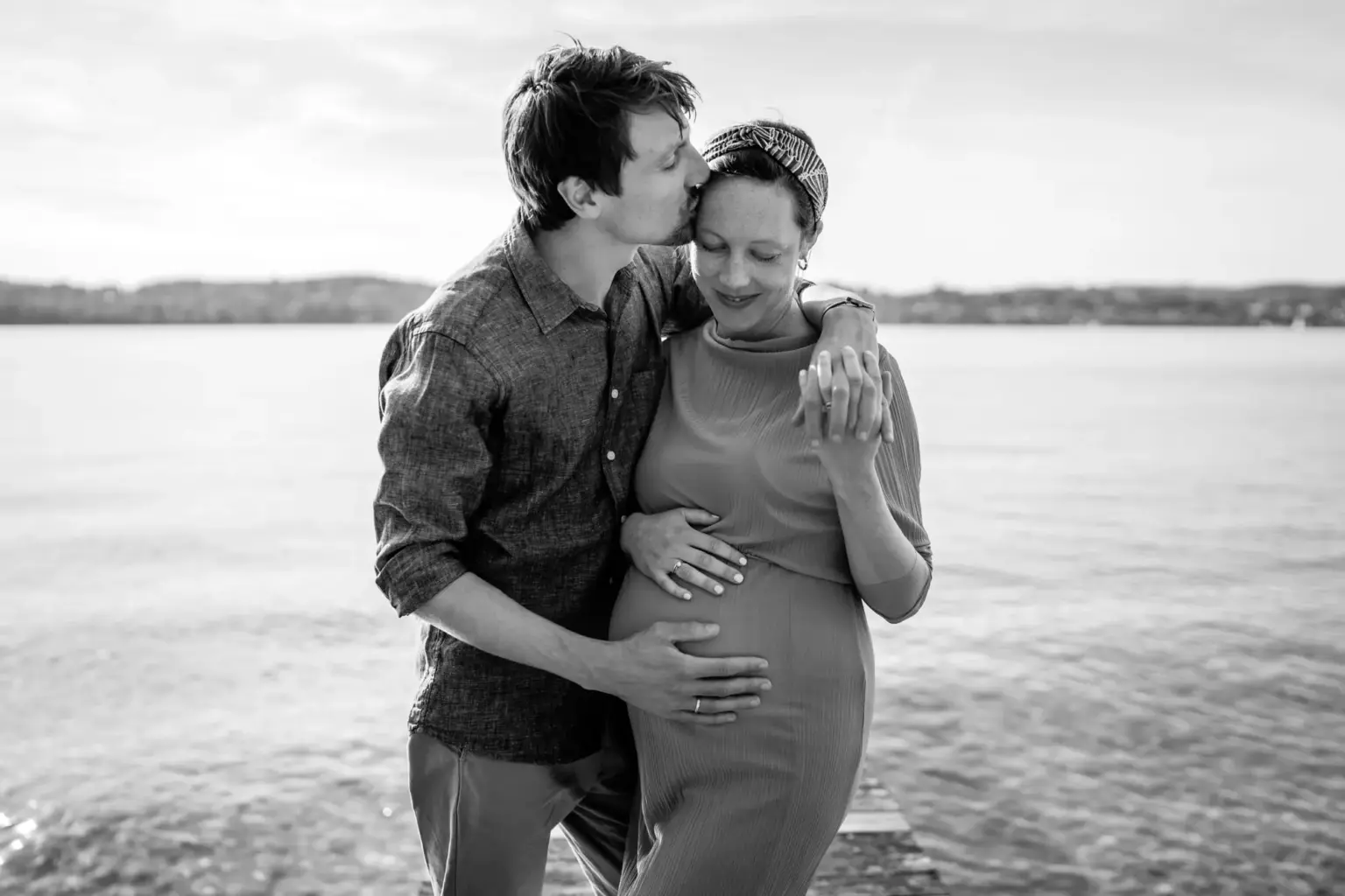 Paar in enger Umarmung, Babybauch im Vordergrund, See weich dahinter