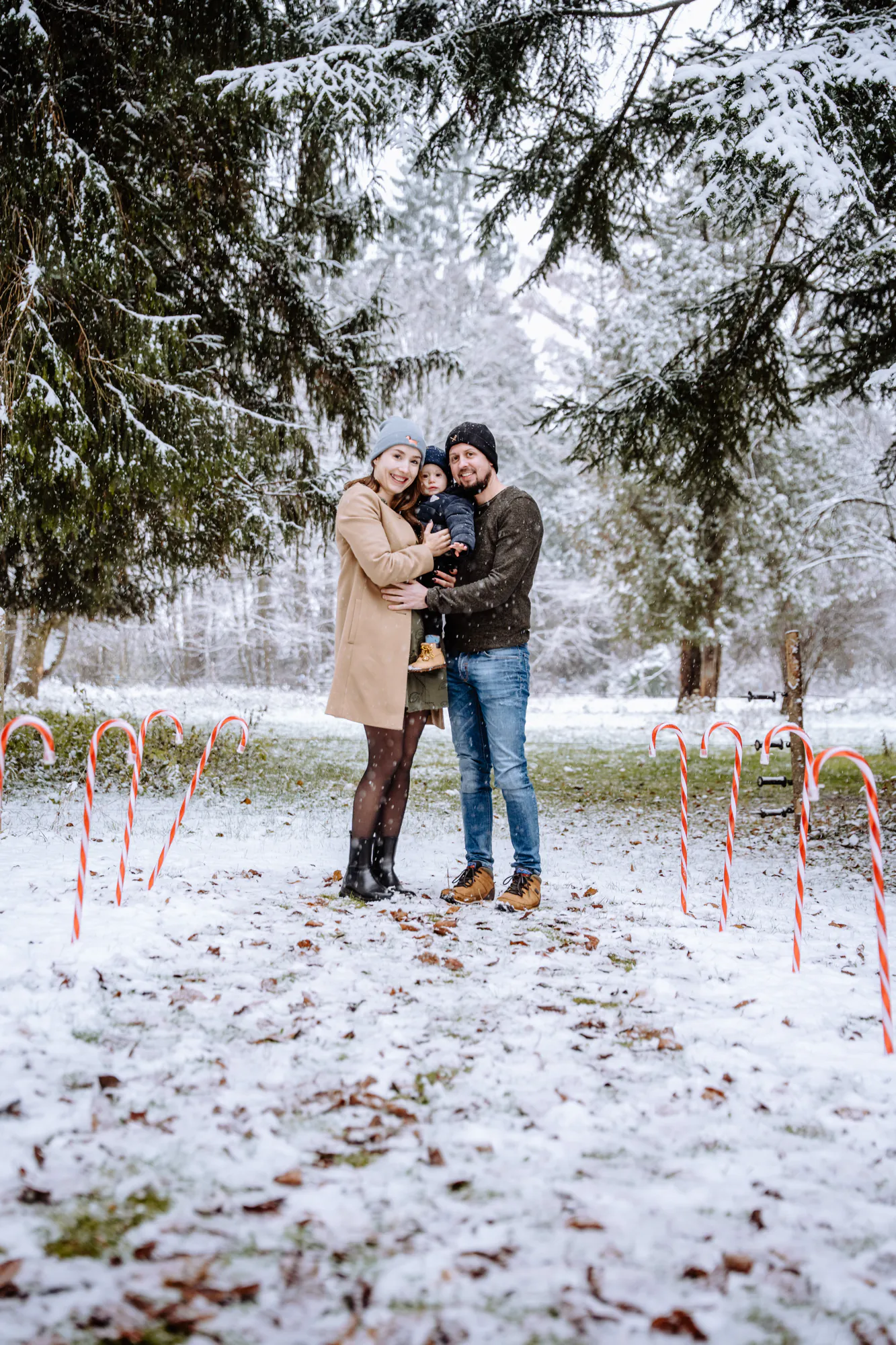 Familie mit Kleinkind steht zwischen Zuckerstangen in einem verschneiten Winterwald