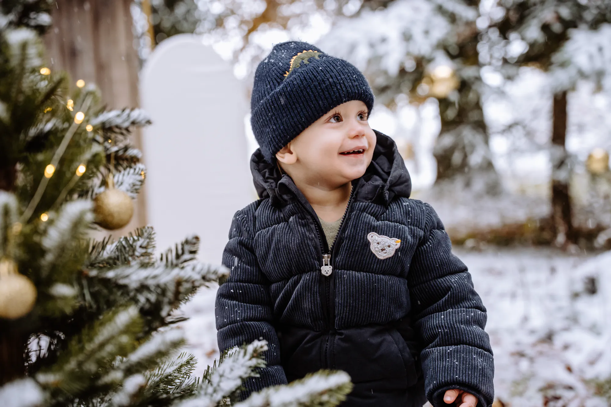 Lachendes Kleinkind mit Wintermütze neben einem geschmückten Tannenbaum im Schnee